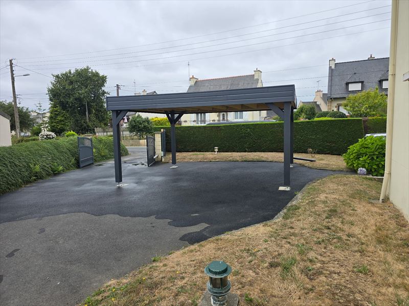 Carport autoportant à quatre poteaux anthracite sur une cour bitumée bordée d'une haie taillée